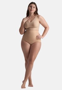 Kurvig kvinna som bär kroppsnära sömlös högmidjad shapewear och matchande bh, stående barfota med en hand på höften och den andra nära nyckelbenet.