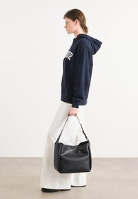 Bolsa de couro preta com um design relaxado e uma alça superior única, combinada com um hoodie azul-marinho e calças largas brancas.