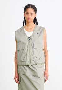 Světlý zelený bezrukávový svetr s předním zipem, dvěma velkými předními kapsami a hladkým povrchem. Nosený přes krémový top s logem.