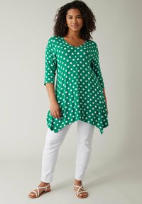 Grøn tunika med polka prikker, V-udskæring og trekvartærmer, parret med hvide slim-fit bukser og sandaler med remme.