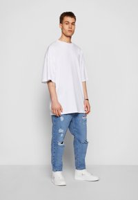 T-shirt blanc oversize à manches courtes, associé à un jean bleu clair déchiré avec des trous, et des baskets blanches.