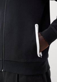 Veste noire zippée avec poche zippée bordée de blanc, main droite partiellement dans la poche, poignets et ourlet côtelés visibles.