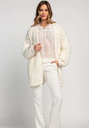 Gruba, biała sweterowa kardiganka z teksturowanym, kablowym wzorem. W zestawie z lekkim białym topem i dopasowanymi białymi spodniami.