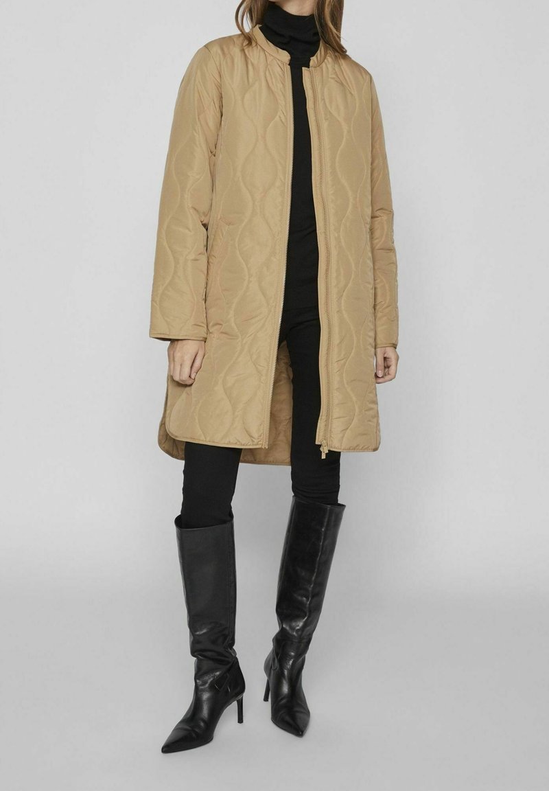 Manteau matelassé beige avec col montant, fermeture éclair et manches longues, associé à un pantalon noir ajusté et des bottes en cuir montantes jusqu'aux genoux.