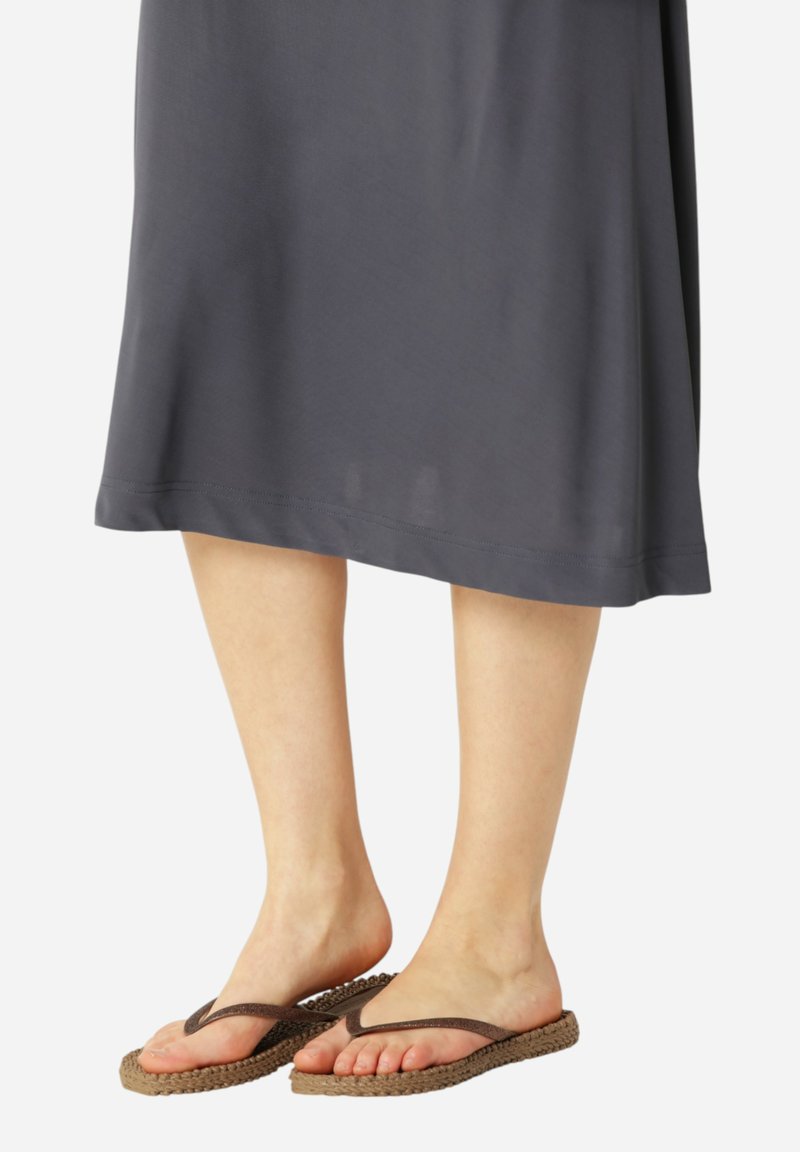 Nohy oblečené v tmavě šedé midi sukni a hnědých wovinových žabkách s tenkými popruhy, stojící na světlém hladkém povrchu.