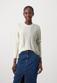 Cremfarbener Strickpullover mit einem strukturierten Chevron-Muster, rundem Ausschnitt und gerippten Bündchen, kombiniert mit einem blauen Jeansrock mit Schlitz.
