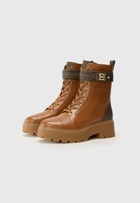 Bottines en cuir marron avec une sangle texturée marron, ornée d'un logo doré, et une semelle en caoutchouc épaisse avec un bord denté.