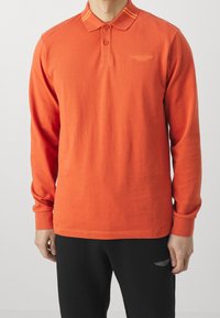 Pitkähihaista polo-paitaa kirkasta oranssia puuvillaa, jossa on keltaraitainen kaulus, kolme napinaukkoa ja logo rinnassa.