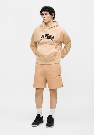 Jeune homme aux cheveux bouclés portant un sweat à capuche beige "BARROW" et un short assorti, des chaussettes blanches et des baskets noires, debout les mains derrière le dos.