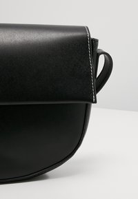 Borsa a tracolla in pelle nera con forma tondeggiante, texture liscia e cuciture bianche in contrasto sulla chiusura a patta.