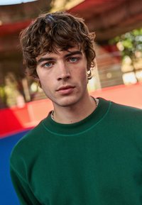 Groene sweatshirt met een ronde halslijn, textuurstof en subtiele stiksels. De achtergrond bevat rode en blauwe kleuren.