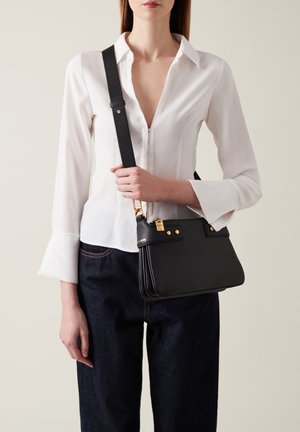 Borsa a tracolla nera in pelle con hardware dorato, forma strutturata, strapola staccabile e due manici superiori, indossata a tracolla con una blusa bianca.