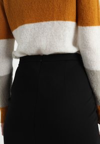Gestrickter Pullover in orange, weiß und grau gestreift, kombiniert mit einem schwarz hoch taillierten Rock. Die Textur wirkt weich und flauschig.