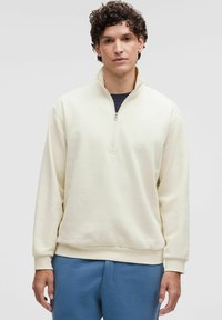Felpa beige chiaro con zip a quarto e maniche lunghe, abbinata a pantaloni blu. Il tessuto appare morbido con una superficie texturizzata.