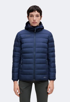 Persona con chaqueta acolchada con capucha azul marino, cremallera frontal y logo en el pecho, combinada con pantalones oscuros sobre un fondo liso.