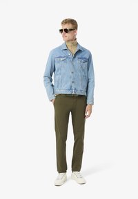 Giacca di jeans azzurra, dolcevita beige, pantaloni verde oliva e sneakers bianche. Modello che indossa occhiali da sole verdi, in piedi in modo informale.