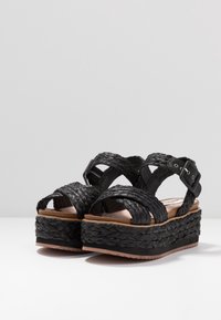 Svarta vävda plattformsandaler med breda remmar, spänne och en texturerad sula. Höjd korkliknande bas ger stabilitet.