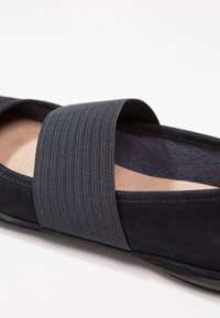 Gros plan d’une chaussure noire à enfiler avec une large bande élastique texturée sur le dessus et une semelle intérieure coussinée couleur fauve.