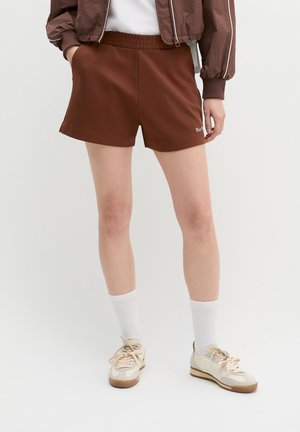Person iført brune shorts med elastisk talje, hvide sokker, cremefarvede sneakers og en brun jakke med hænderne delvist i lommerne.