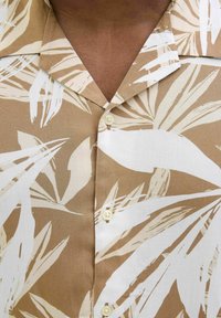 Chemise à manches courtes en beige et blanc, ornée d'un motif de feuilles, avec un col et des boutons. Fabriquée à partir d'un tissu léger avec une texture douce.