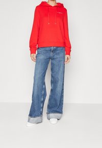Sudadera con capucha roja combinada con jeans de pierna ancha de color azul claro, con puños doblados y zapatillas blancas. Diseño simple con acentos mínimos.
