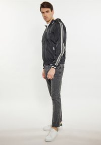 Sort nylon bomberjakke med hvide striber, lynlåslommer og hætte, parret med grå jeans og hvide sneakers. Justeret pasform ved ærmerne.