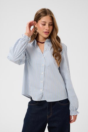 Jeune femme aux longs cheveux portant une chemise rayée bleue et blanche à boutons et un jean foncé, touchant ses cheveux d'une main.