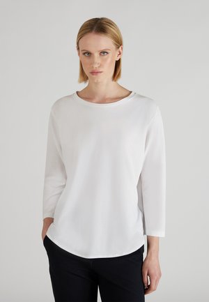 Vrouw met blond haar, gekleed in een eenvoudige witte lange mouw shirt en zwarte broek, staande met één hand in de zak tegen een effen achtergrond.