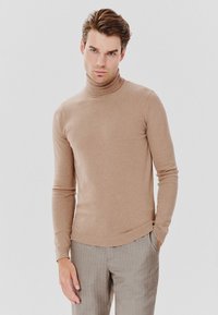 Jasnobrązowy sweter z golfem, wykonany z miękkiej tkaniny. Posiada długie rękawy i dopasowany krój. W połączeniu z szarymi spodniami w prążki.