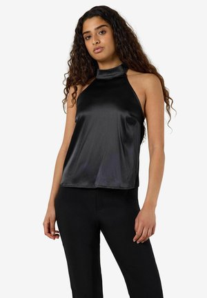 Femme aux longs cheveux bouclés portant un haut noir en satin sans manches à col halter et un pantalon noir, debout devant un fond blanc uni.