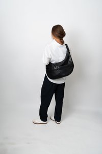 Mulher com cabelo trançado está de costas, vestindo uma camisa branca, calças pretas com riscas, sapatilhas brancas e uma grande bolsa preta de ombro.