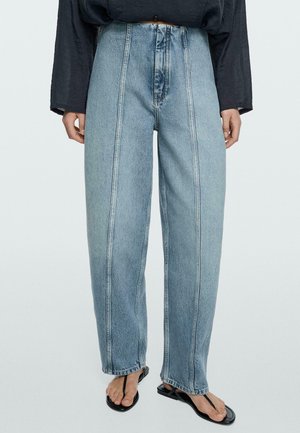 Osoba nosząca jasnoniebieskie dżinsy z wysokim stanem i pionowymi szwami, czarny top z długim rękawem oraz czarne sandały thong stoi na jednolitym tle.