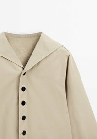 Veste chemise beige avec un large col, six boutons noirs sur le devant et un tissu lisse, légèrement texturé.