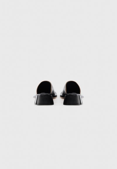 Une paire de mules noires en cuir à talons bas avec des orteils carrés, vues de l'arrière sur un fond blanc uni.