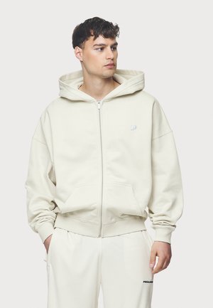 Veste à capuche zippée beige clair en tissu doux, avec une capuche, des poches kangourou et des poignets côtelés. Petit logo sur la poitrine gauche.