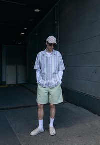Cămașă cu mânecă scurtă, cu dungi, pantaloni scurți verzi deschis, șapcă bej, ochelari de soare, sosete albe și pantofi slip-on casual, pe un fundal de perete gri.