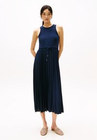 Rochie plisată de culoare bleumarin, cu design fără mâneci, având talie cu șnur și o siluetă vaporos, asortată cu pantofi tip slip-on din împletitură.