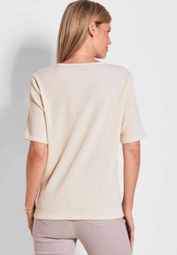 Helles beigefarbenes Kurzarmtop mit strukturiertem Rippenmuster, rundem Halsausschnitt und lockerer Passform, von hinten gezeigt.