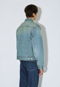 Veste en jean bleu clair avec une finition légèrement délavée, col classique et deux boutons à la taille. Portée avec un jean bleu foncé.