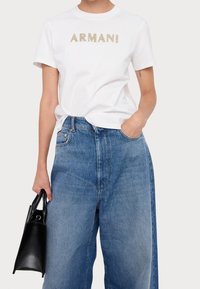 Vit bomullst-shirt med "ARMANI" i guldfärgade bokstäver, kombinerad med ljusblå jeans med vida ben. Svart läderhandväska hålls i ena handen.