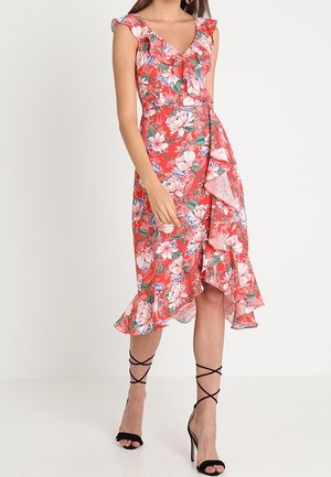 Vrouw draagt een mouwloze rode bloemen wikkeljurk met ruches en zwarte hoge hakken met bandjes, staand tegen een effen witte achtergrond.