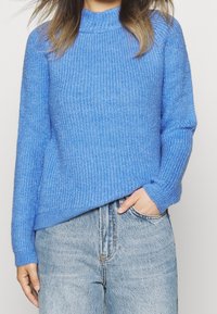 Pull bleu tricoté avec une texture côtelée, col montant et manches longues, associé à un jean en denim bleu clair.