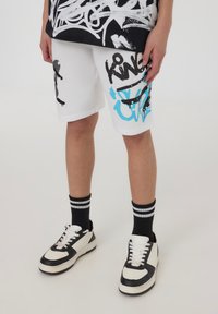 Pantaloni corti bianchi con design in stile graffiti neri e blu, abbinati a scarpe da ginnastica bianco e nere e calzini a righe nere.