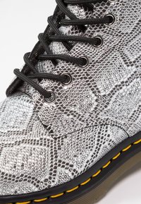 Gros plan sur une botte argentée à motif peau de serpent avec des lacets noirs et des coutures jaunes sur la semelle noire.