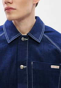 Denimjakke i mørkeblå med hvite sømmer, har en spiss krage, sølvfargede knapper og en brystlomme med en logoetikett.