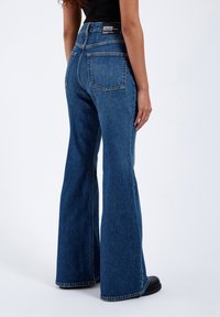 Flared denim jeans in medium blue, featuring a high waist, two back pockets, and a clean hem. Made of rigid denim fabric.