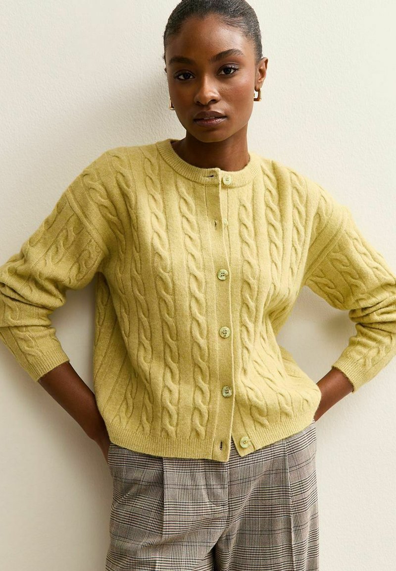 Cardigan in maglia di colore giallo limone con motivo a trecce, bottoni verdi e polsini a coste. Abbinato a pantaloni a quadri per un look stratificato.