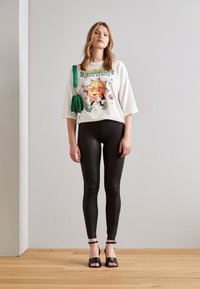 T-shirt graphique blanc avec un design de soleil coloré, leggings noirs à motif serpent, talons fendus noirs et un sac à main vert.