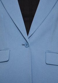 Svetle modrý blazer s hladkým povrchem, s klopami, zapínání na jeden knoflík a dvěma předními kapsami. Pozadí je černé.