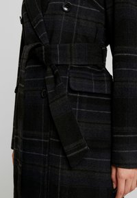 Cappotto di lana a scacchi scuri con cintura larga legata in vita, tasca con patta e grandi bottoni neri, indossato sul torso e sul braccio di una persona.